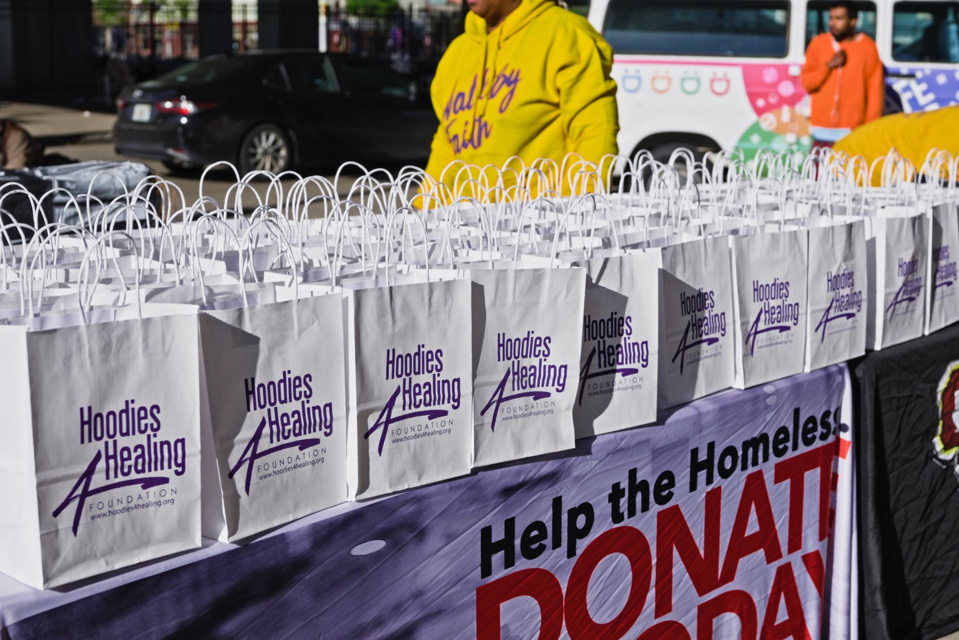 A man in a yellow hoodie stands in front of a table full of bags that say hoodies healing