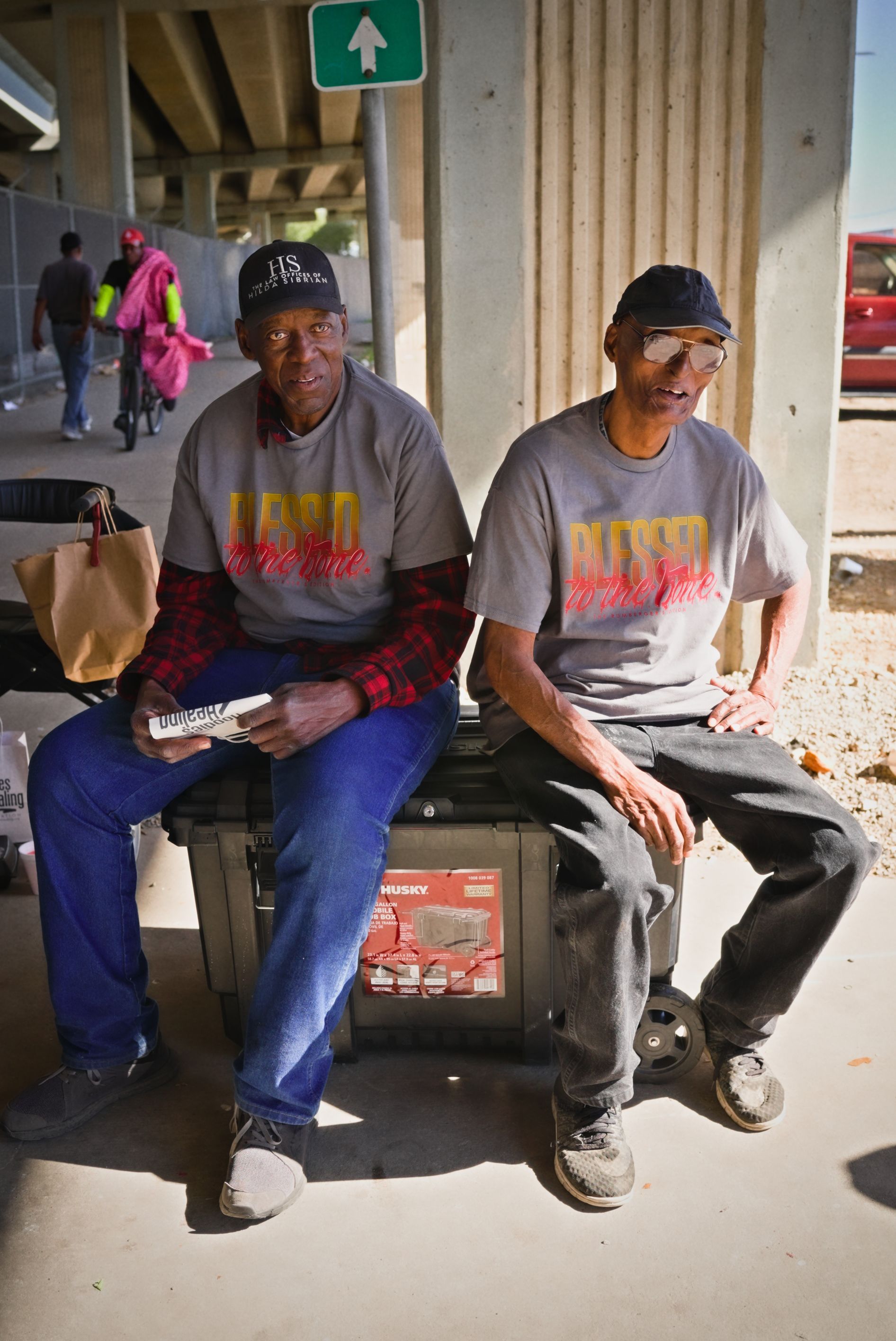 Two men are sitting next to each other on a bench under a bridge.