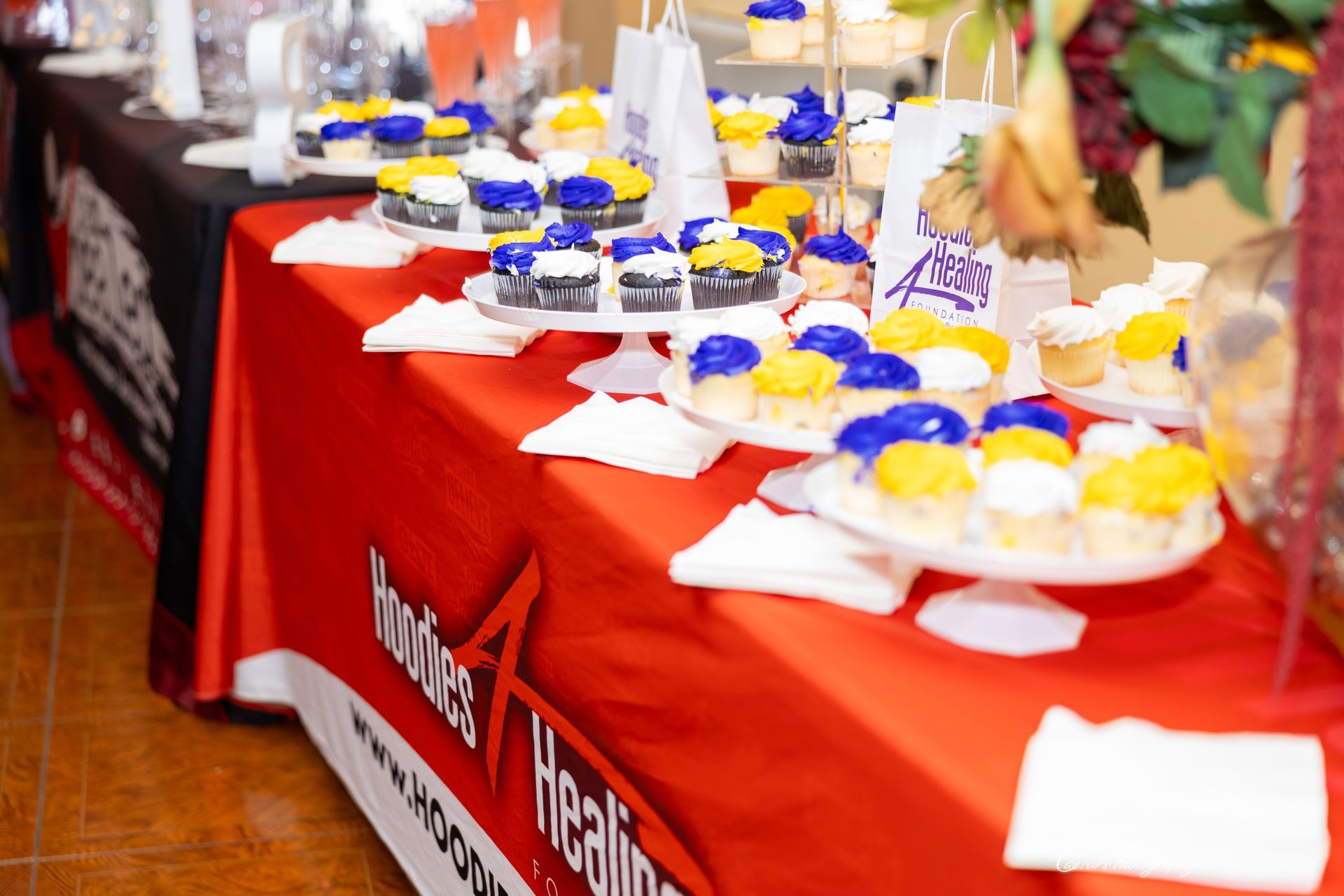 A table with a red table cloth and cupcakes on it.