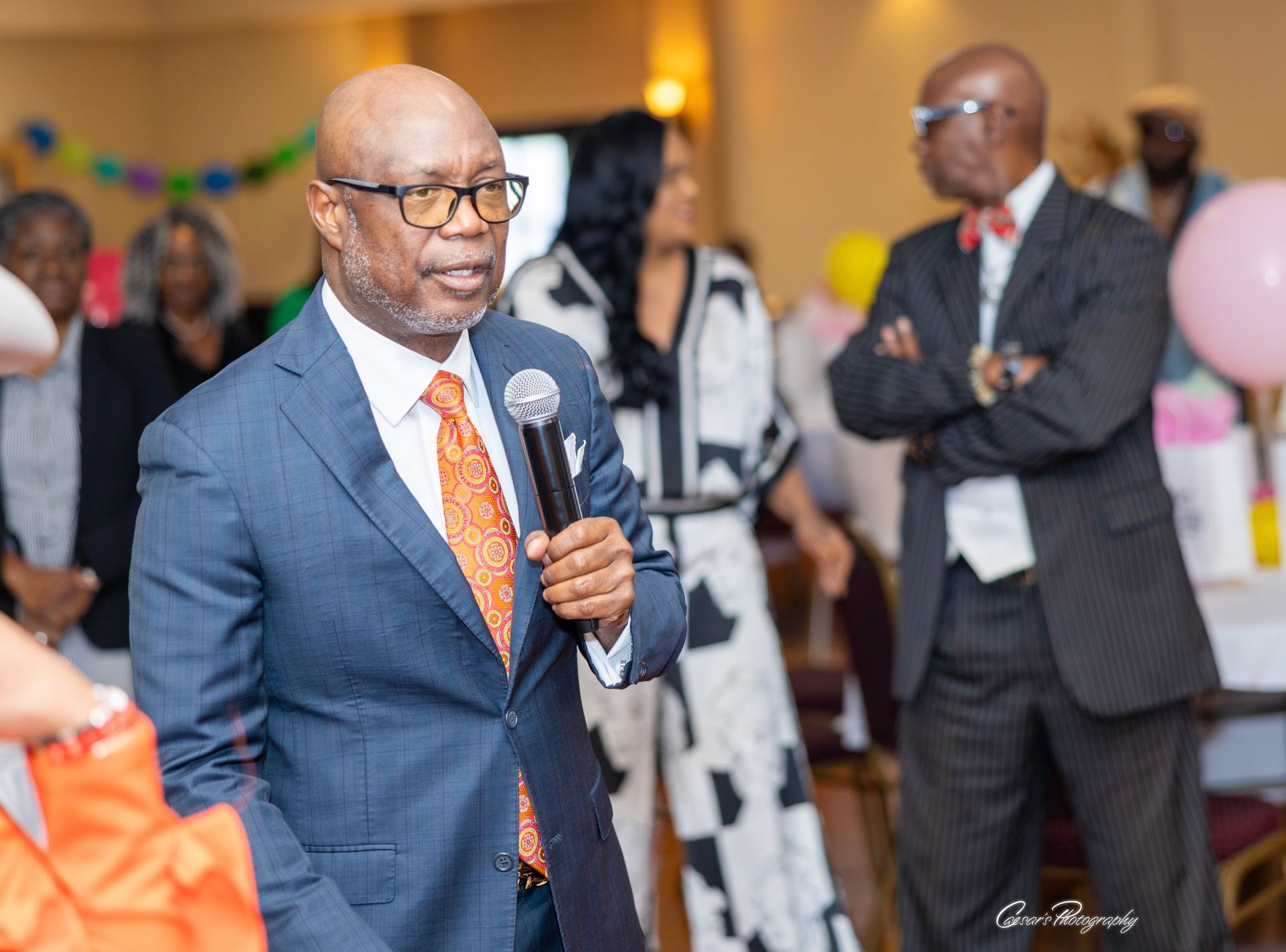 A man in a suit and tie is speaking into a microphone at a party.