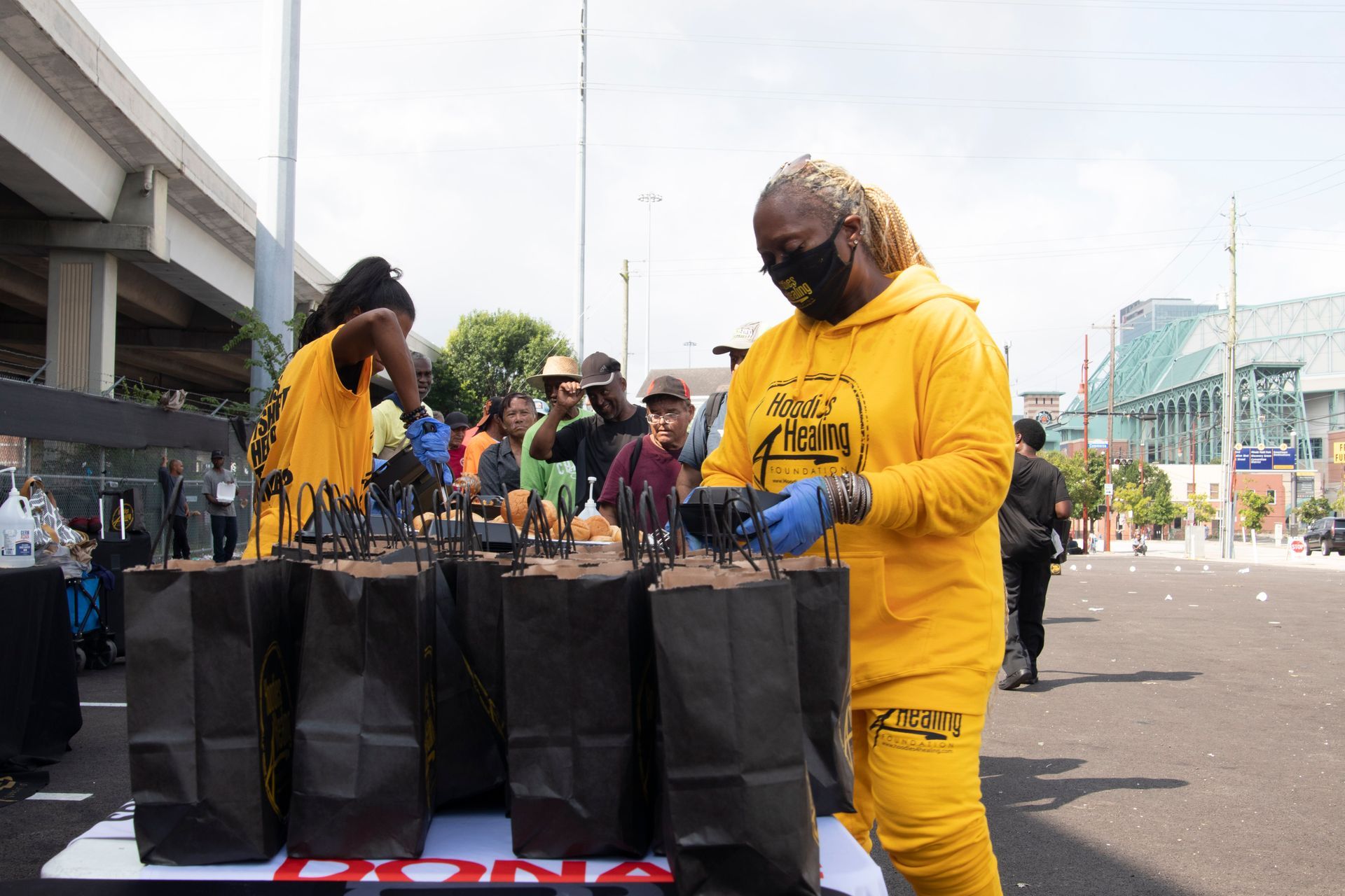 A woman in a yellow hoodie is standing in front of a table full of bags.