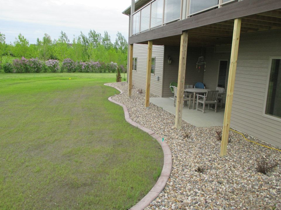 A grassy backyard with a rock border and a covered patio under a house with a glass balcony.