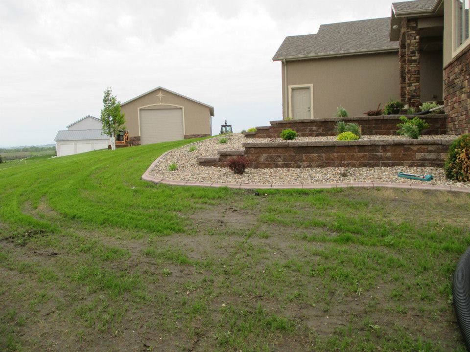 A house with a tan exterior and a retaining wall, a garage, and a green lawn on a cloudy day.
