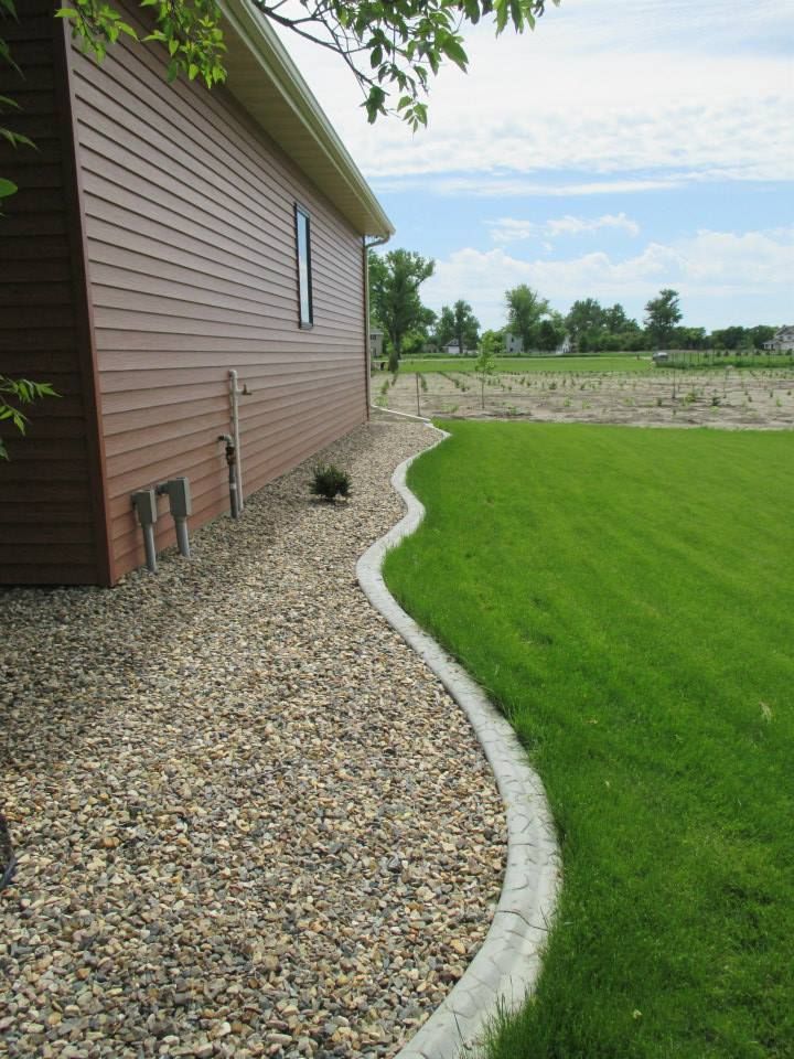 Brown-sided house with a gravel bed and a concrete lawn edge, next to green grass under a blue sky.
