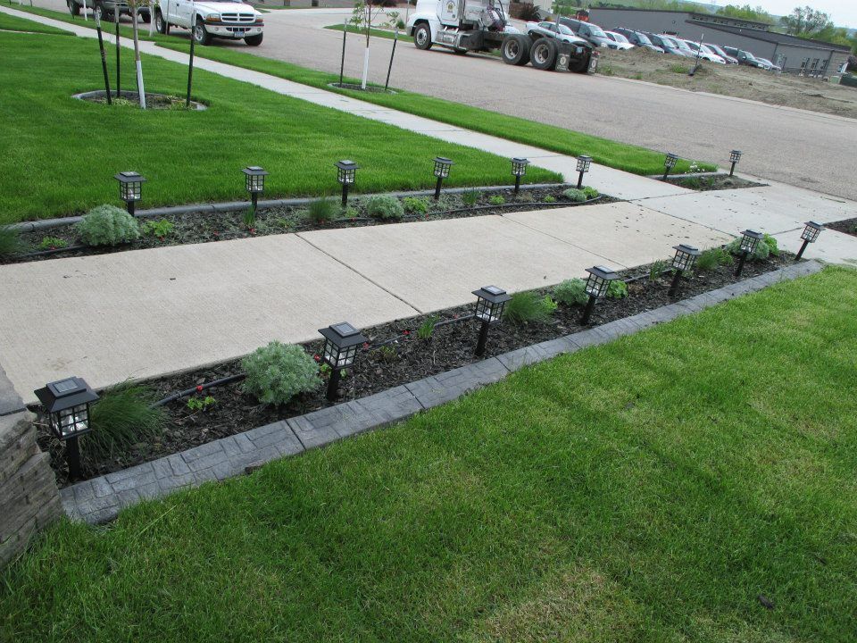 Concrete walkway with solar lights and landscaping in a residential area. Green grass and gray paving stones.