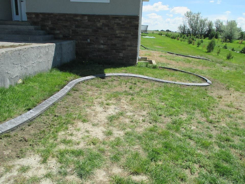 Gray stone edging curves around a house foundation and lawn with patchy grass on a sunny day.