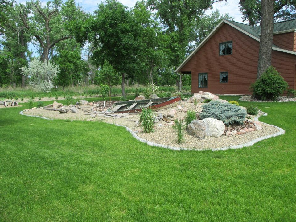 A house with brown siding sits on a grassy lawn with a rock garden. Trees and shrubs surround the property.