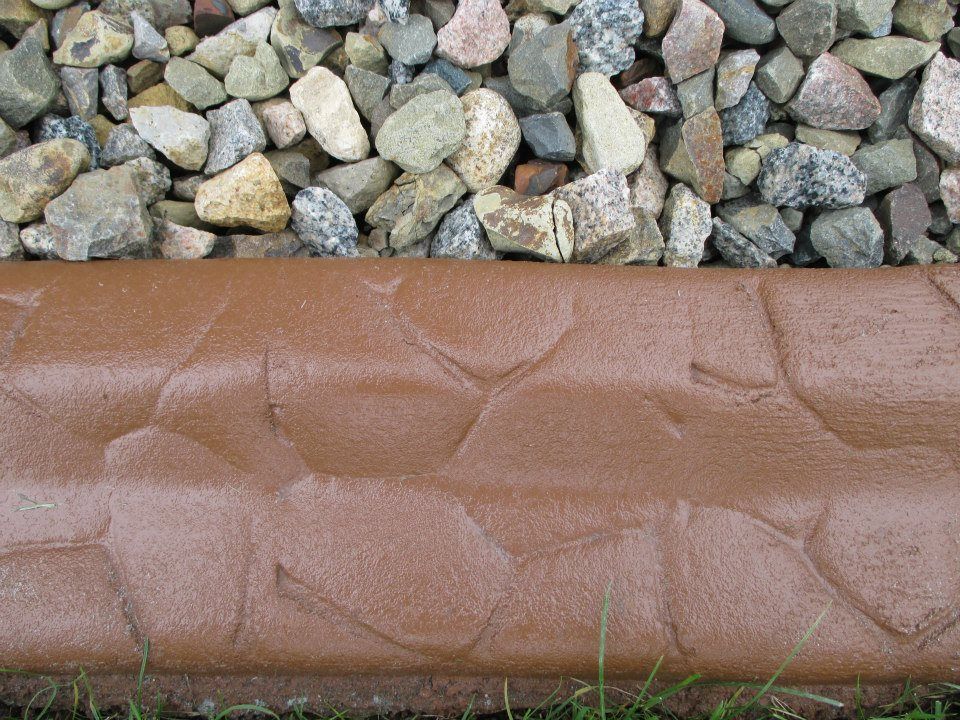 Brown stone-like edging separates a bed of small, multicolored gravel from green grass.