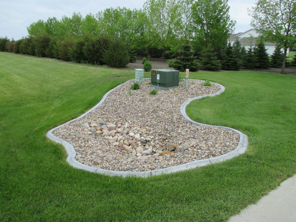Rock-filled landscaping around an electrical box in a grassy yard; trees in the background.