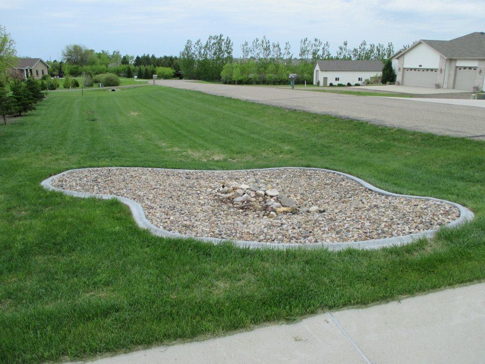 Rock garden with gray border in a grassy front yard, residential neighborhood in background.