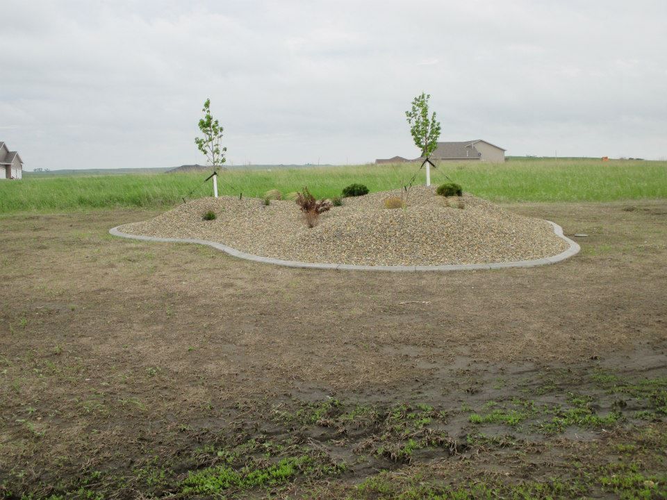 Gravel landscaping bed with small trees and plants, bordered by concrete on a grassy lawn, with a cloudy sky.
