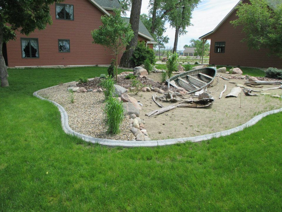 Landscaped garden bed with boat, rocks, and small plants, bordered by concrete curb, near brown buildings.