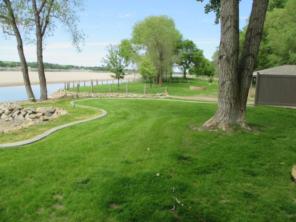 Grassy park next to a river, with trees, rocks, and a brown building. Bright sunny day.
