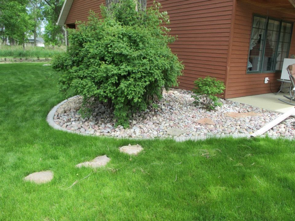 Stone bed with green shrub and small tree next to a brown house. Stepping stones on the grass.