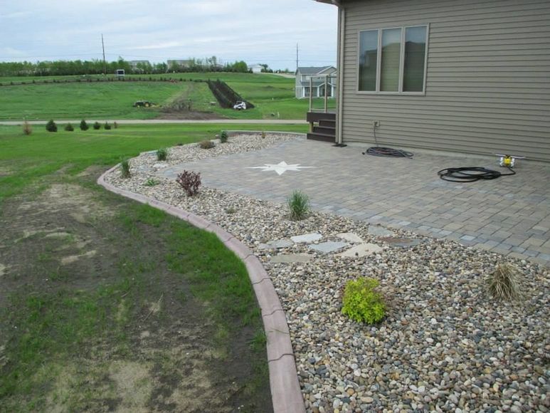 Patio with stone pavers and rock garden next to a tan house. Green grass surrounds the patio.