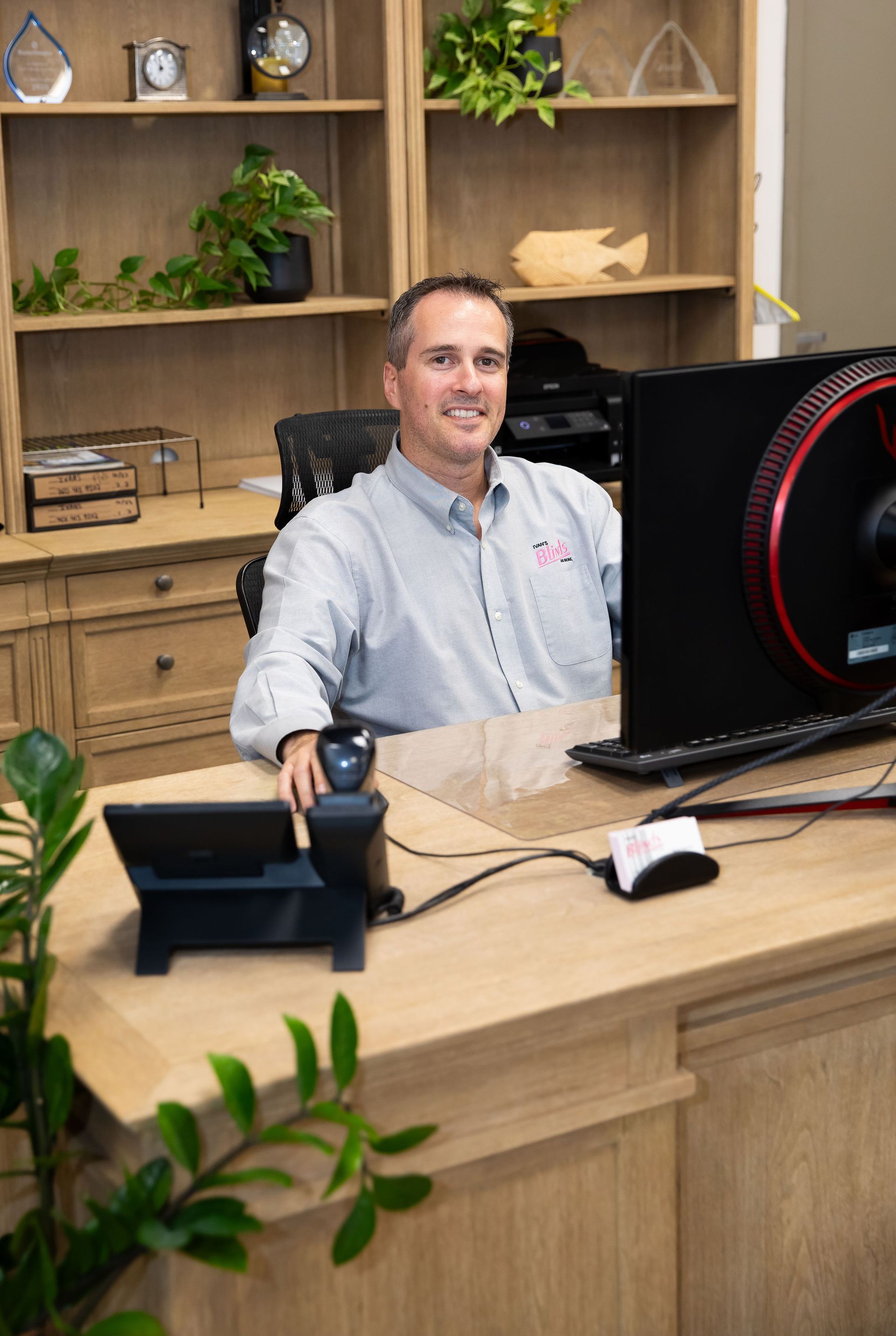 Man smiling at desk, working on computer. Office setting with wooden shelves and desk.