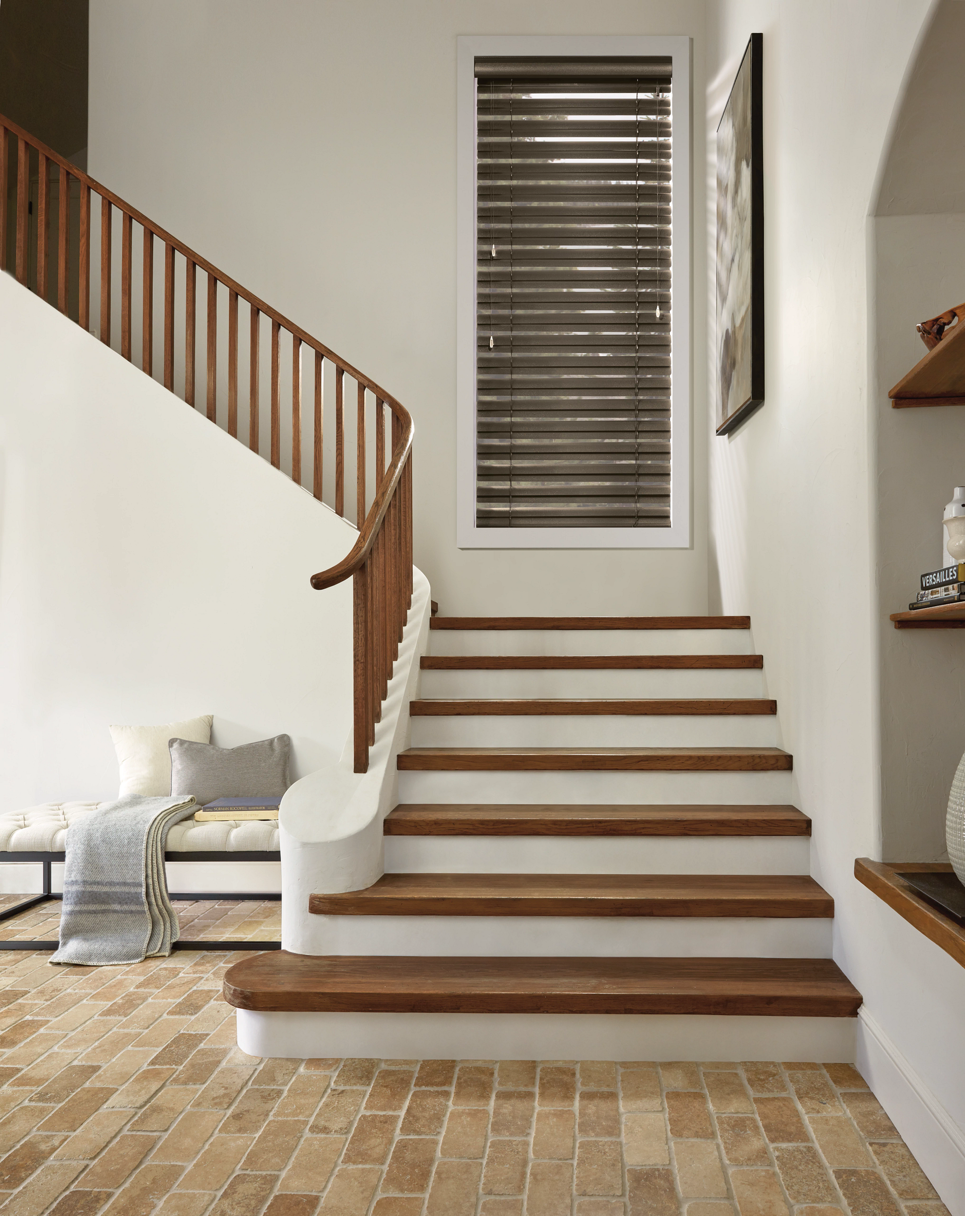 Staircase with wooden steps and railing, brick floor. Window with blinds and shelves on wall.