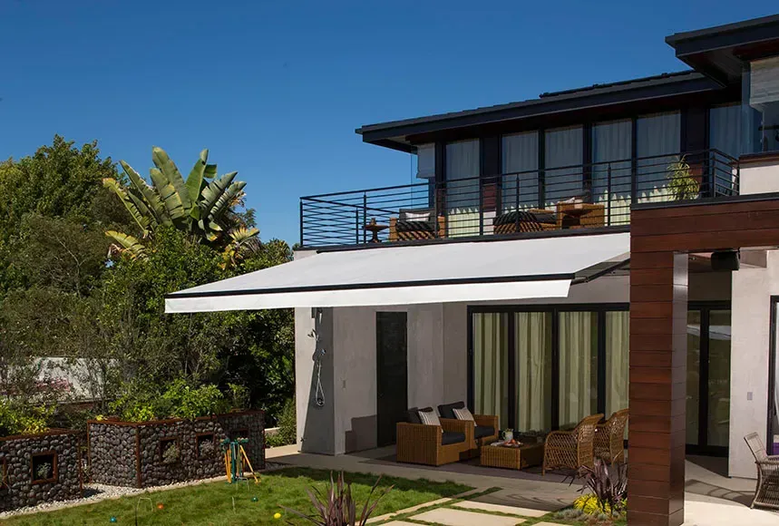 Modern house exterior with retractable awning and balcony. Green lawn, blue sky, and lush vegetation.