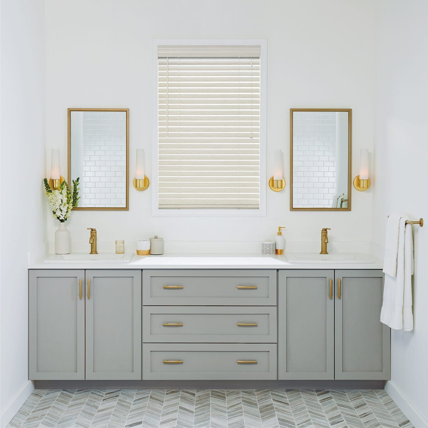 Gray bathroom vanity with gold accents, mirrors, and window blinds. White walls, patterned floor.