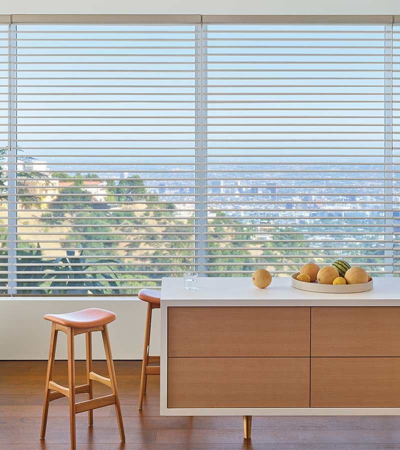 Kitchen with wood cabinetry, a white countertop, sheer shades, bar stools, and horizontal blinds overlooking a cityscape.