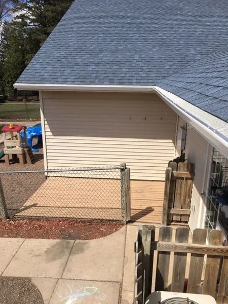 A beige-sided building corner with a wooden deck and chain-link fence, viewed from a concrete walkway.