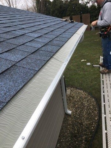 A worker on scaffolding installs white gutter covers on a residential roof with blue asphalt shingles.