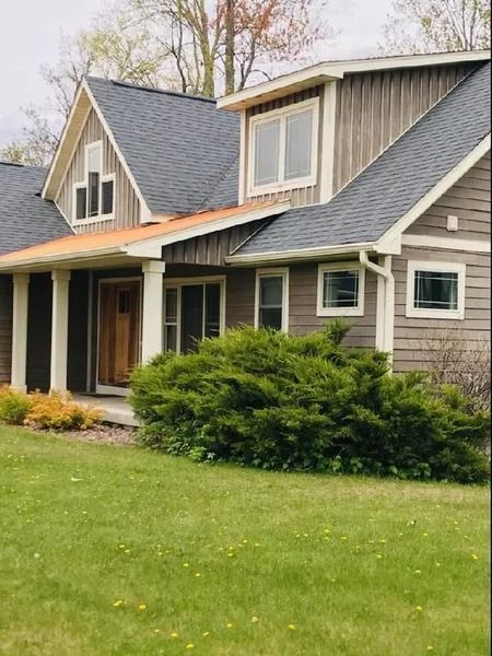 A multi-story house with gray wood siding, a dark shingled roof, a covered porch, and a large green bush in the front yard.