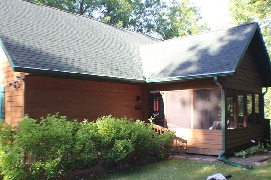 A wooden cabin with a dark roof and a screened-in porch, surrounded by green bushes and trees on a sunny day.