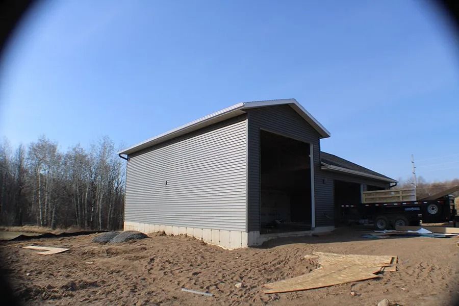 A metal-sided building under construction with open garage bays, situated on a dirt lot against a clear blue sky.