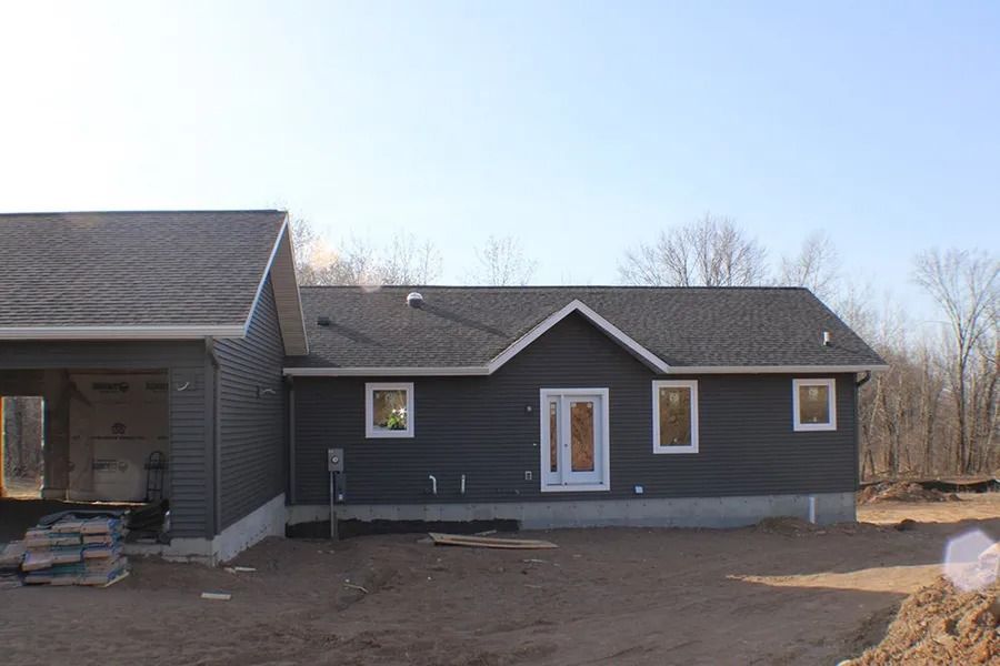 A new single-story house with dark grey horizontal siding, a gabled roof, and a white front door, set on a dirt lot.