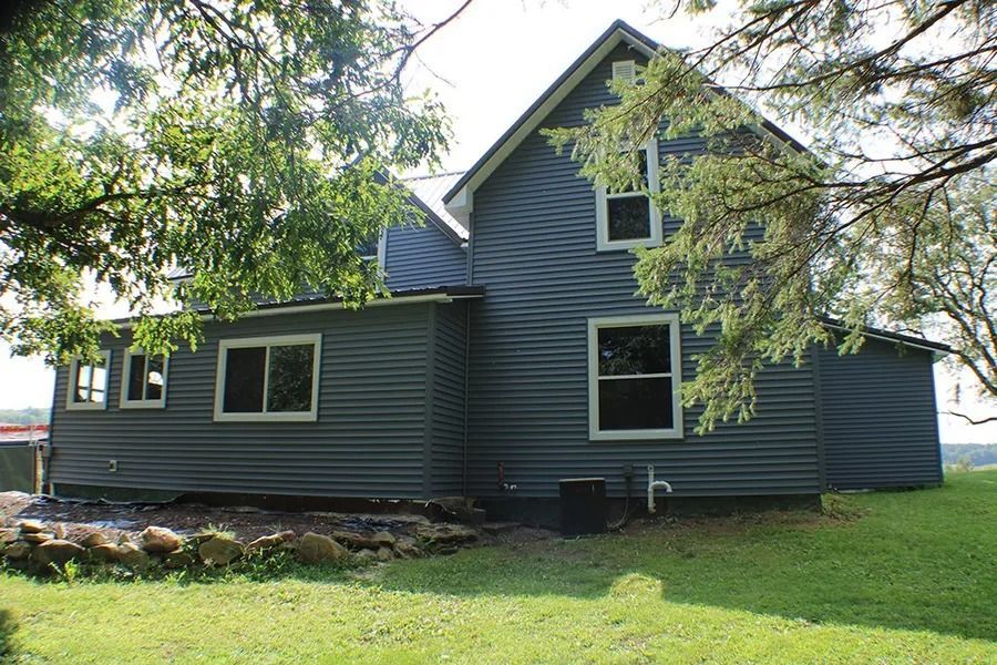 A two-story house with dark blue siding sits on a grassy lawn surrounded by trees.