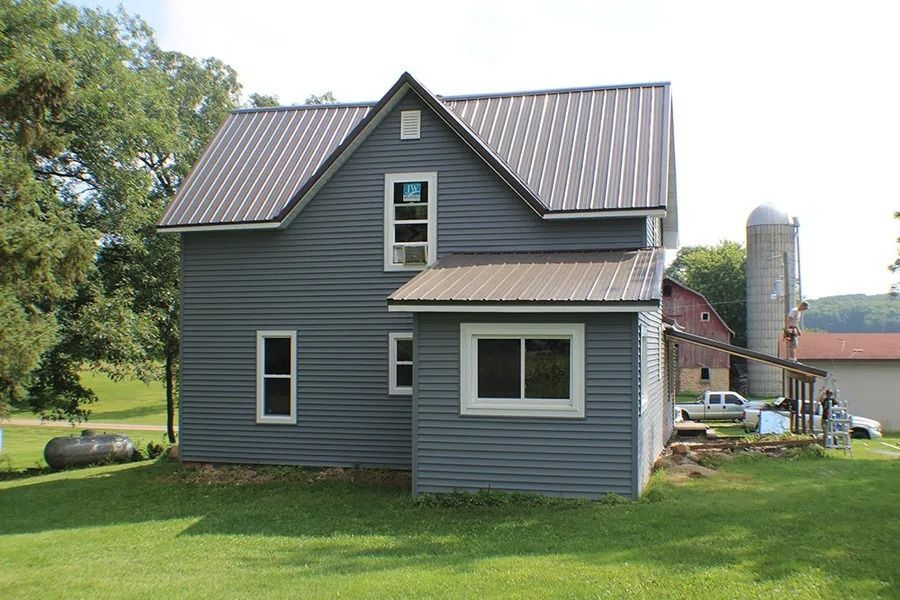 A blue two-story house with a metal roof, located in a rural setting with a silo and farm buildings in the background.