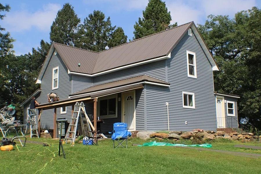 A blue farmhouse with a metal roof under renovation, featuring scaffolding, a ladder, and tools on a grassy lawn.