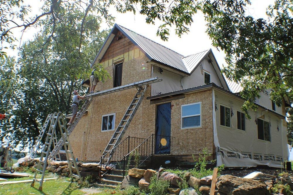 Two workers on ladders renovate the exterior of a two-story wooden house with a metal roof surrounded by trees.
