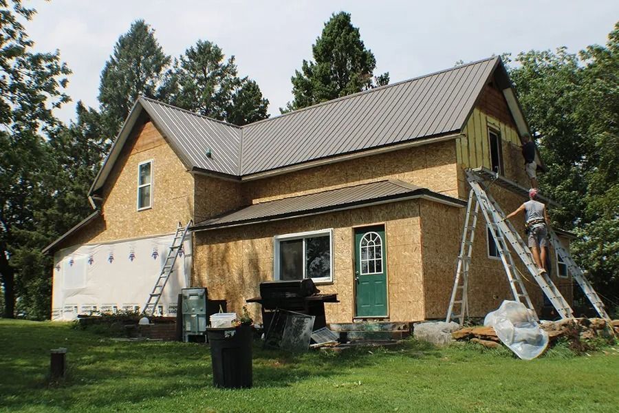 A person works on the exterior of an unfinished house with wooden siding, a metal roof, and a green door in a rural area.