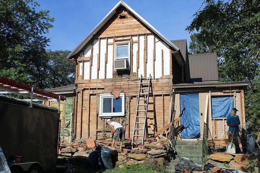 Two people work on a house exterior stripped to its wooden frame, with rubble at the base and a ladder leaning against it.
