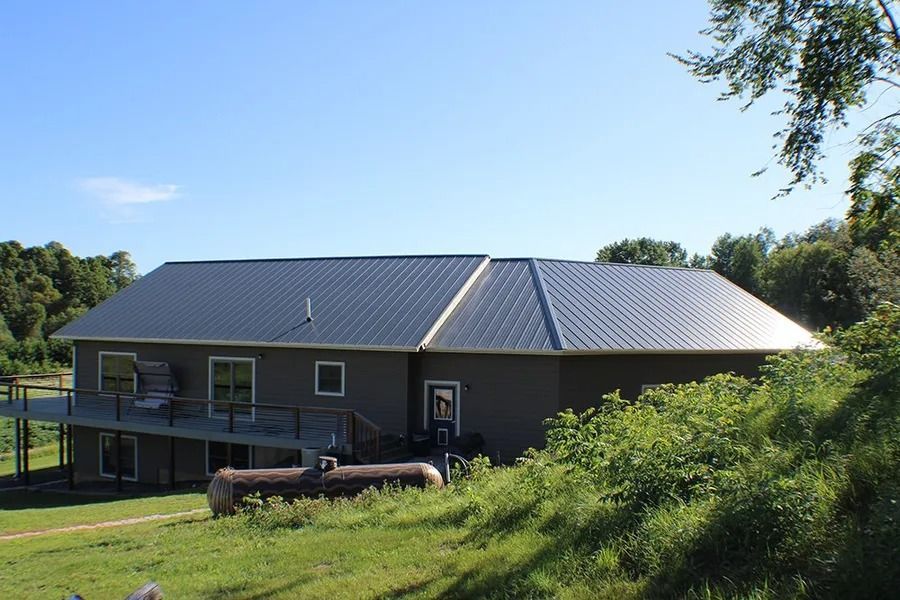A gray house with a dark metal roof and a wooden deck, set against a grassy, tree-filled landscape under a clear blue sky.