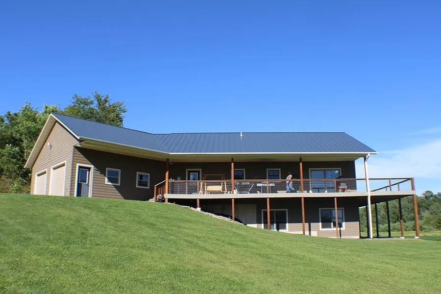 A brown two-story house with a blue metal roof and large wooden deck sits on a grassy hill under a clear blue sky.