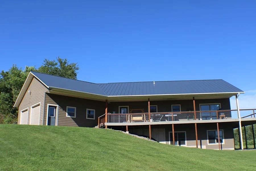 A brown ranch-style house with a blue metal roof and a spacious wooden deck on a grassy hill under a clear blue sky.