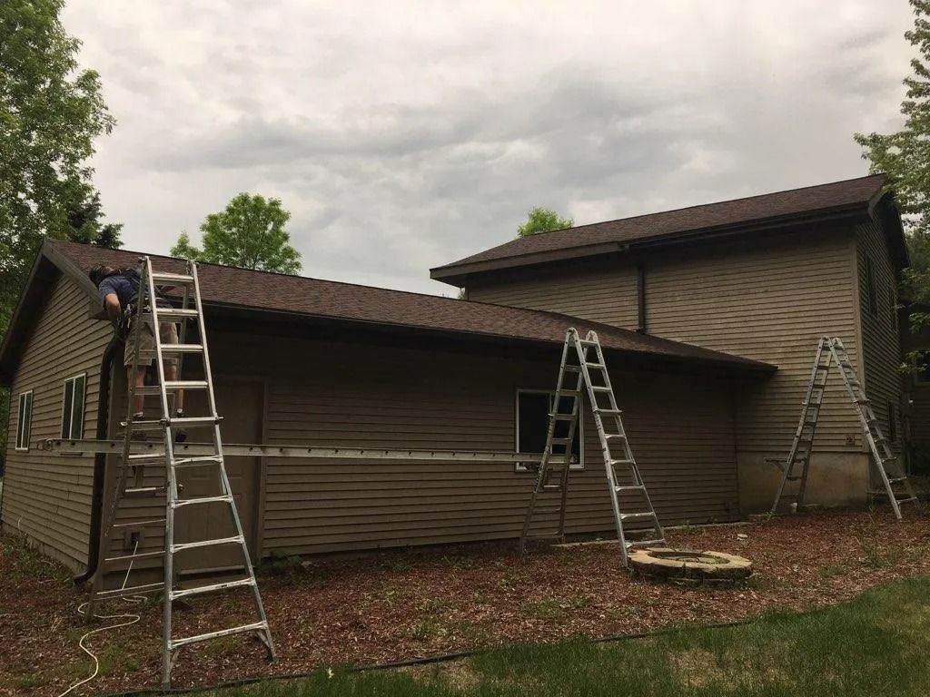 A worker stands on a scaffold supported by two ladders against the side of a brown house during exterior maintenance.