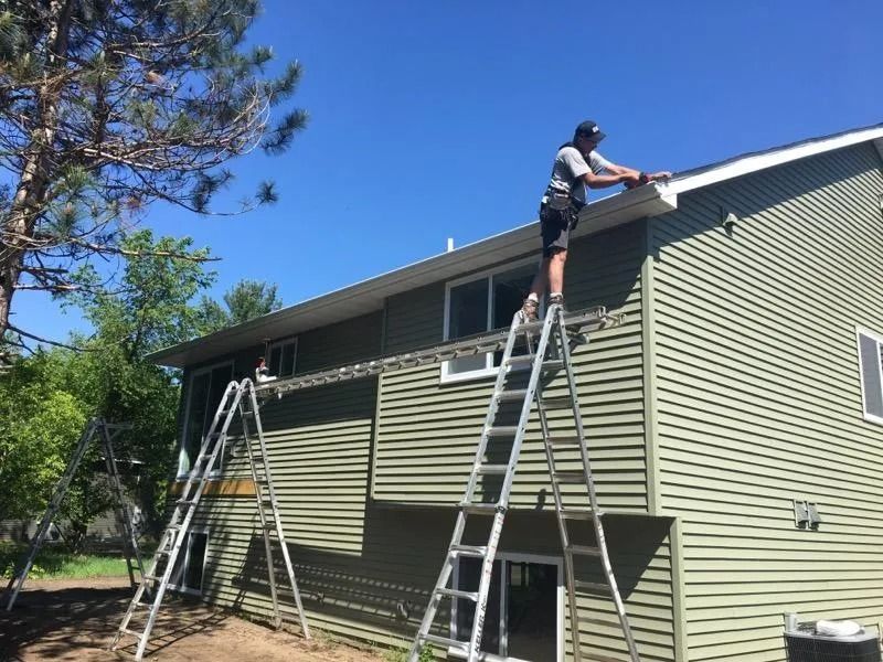 A person on a ladder working on the roofline of a green two-story house.