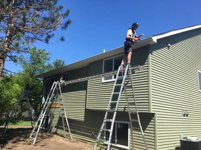 A person works on a house's gutter from a platform supported by two ladders against the green-sided wall.