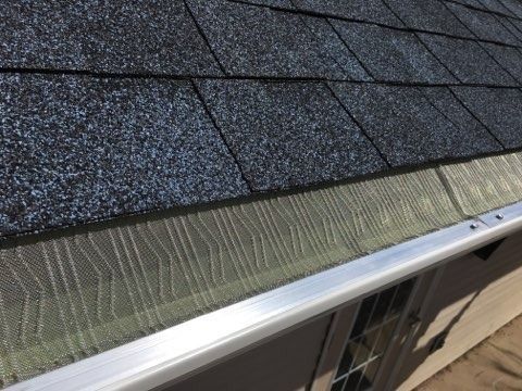A close-up view of a metal mesh gutter guard installed on a house roof with dark gray asphalt shingles.
