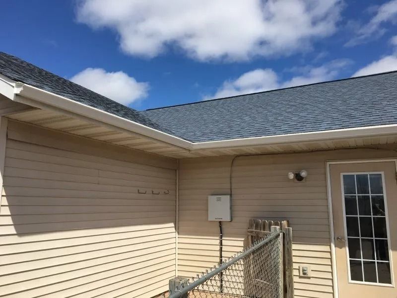 The corner of a tan vinyl-sided house with a grey shingled roof, an exterior door, and a chain-link fence under a blue sky.