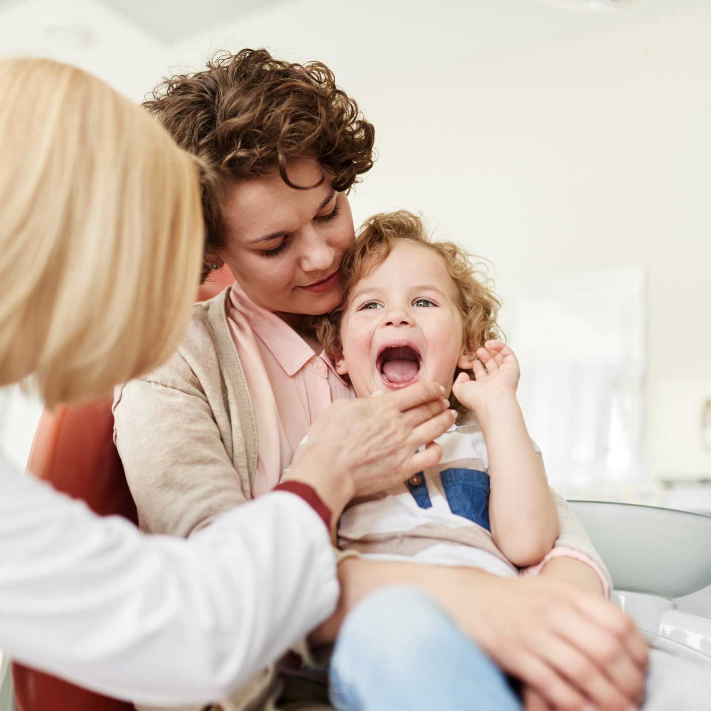 Dentist examining a child's teeth, held by their mother, in a brightly lit clinic.