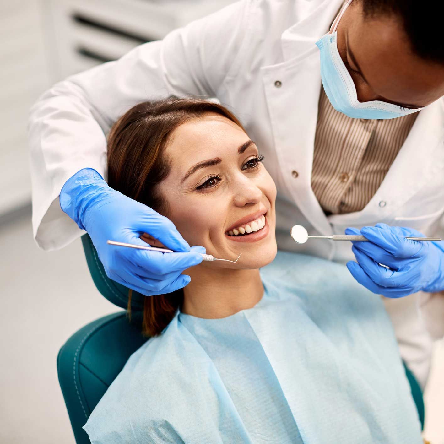 Woman smiling during dental exam; dentist wearing mask and gloves examining teeth.