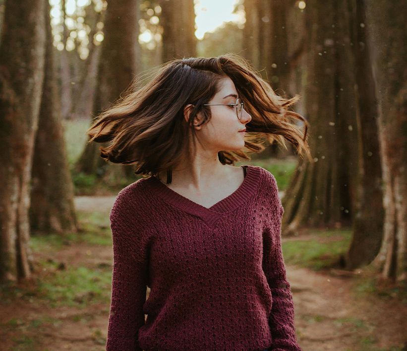 A woman in a purple sweater is standing in the woods with her hair blowing in the wind.