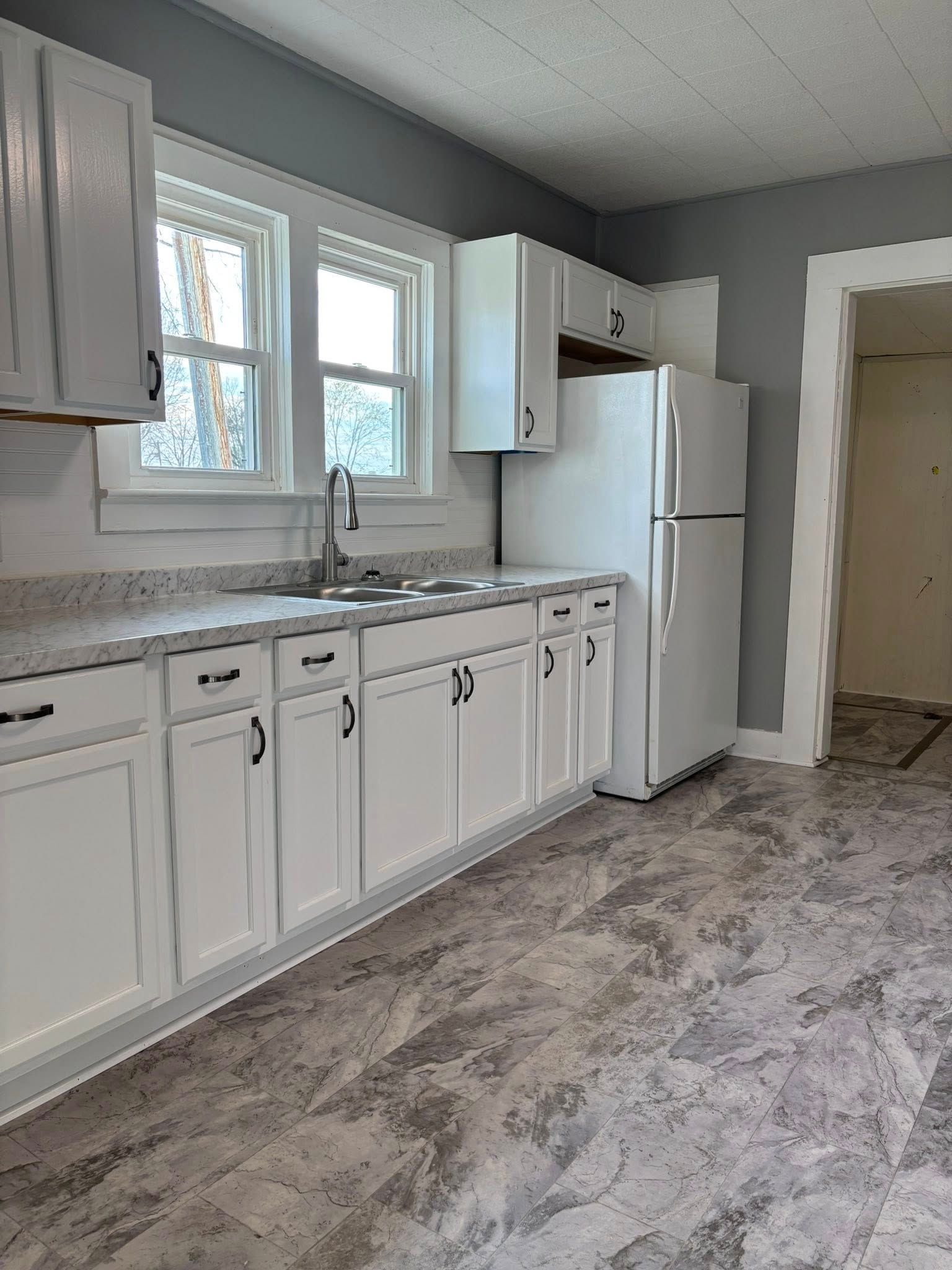 White kitchen with cabinets, sink, and refrigerator, gray walls and flooring, window.
