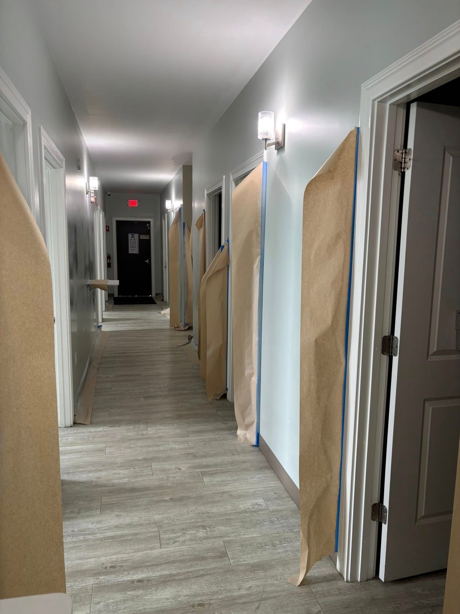 Long hallway with doors and lights. Some doors covered with brown paper. Black door at the end.
