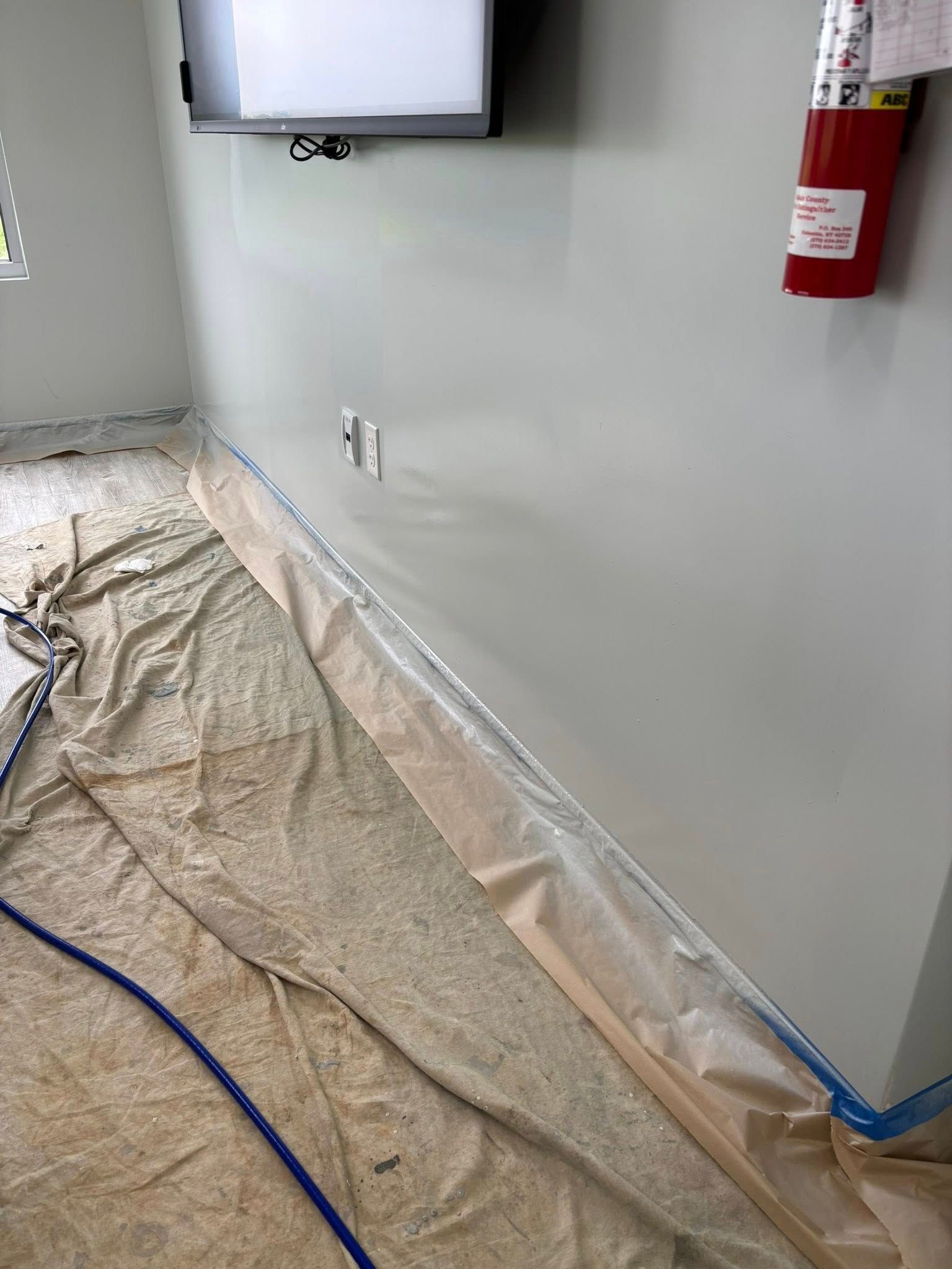 Room with light gray walls; drop cloths protect the floor. Fire extinguisher mounted on wall.